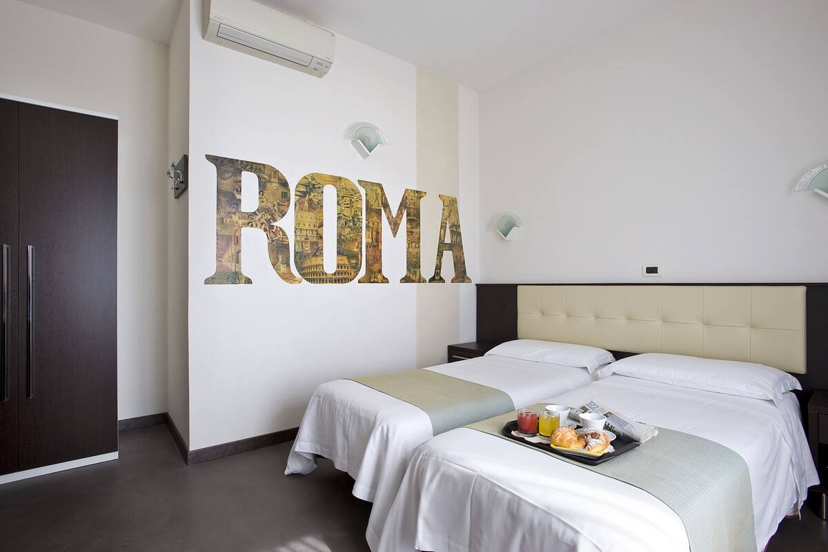 A hotel room with two beds, a large wall decoration spelling "ROMA," and a breakfast tray on the beds.