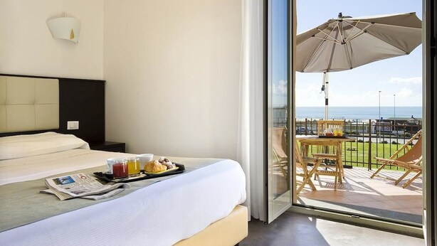 Habitación de hotel con cama, bandeja de desayuno, periódico y balcón con mesa, sillas y sombrilla.