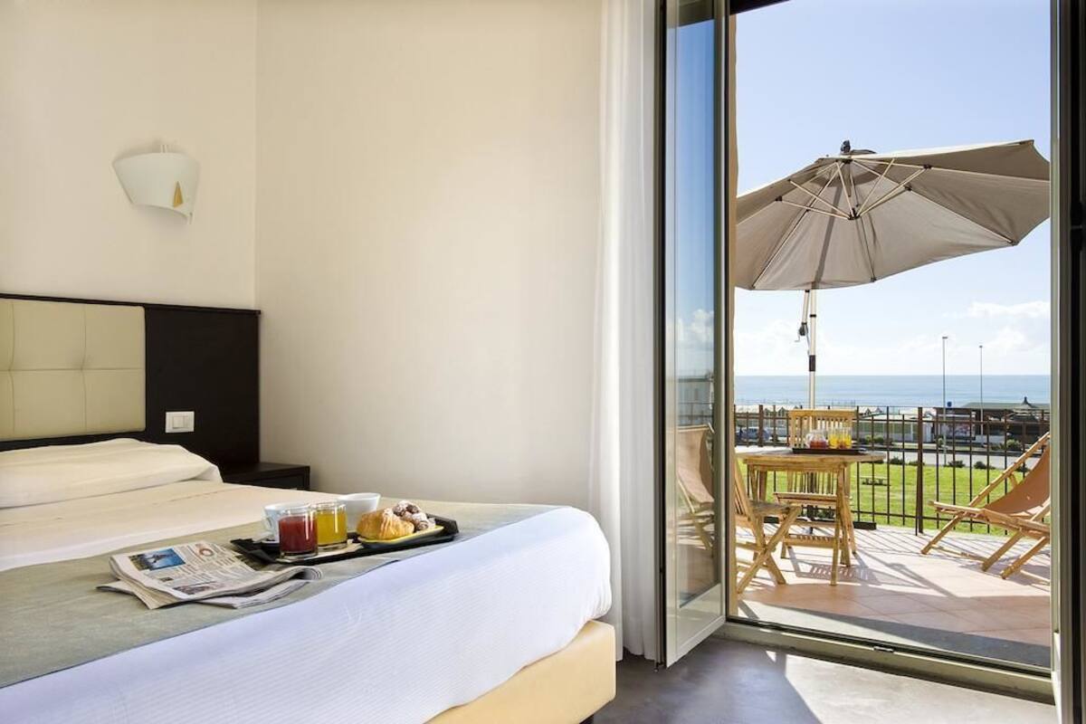 A hotel room with a bed, breakfast tray, newspaper, and a balcony with a table, chairs, and an umbrella.