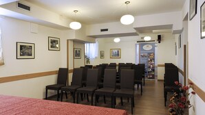 Habitación de hotel con cama, una mesa de comedor y sillas dispuestas para una reunión. Se observan cuadros en las paredes y un cartel en la pared.
