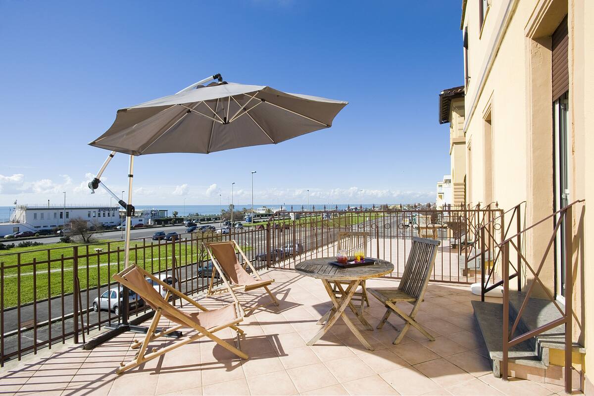 A balcony with a table and chairs, a large umbrella, and a view of the sea and buildings.