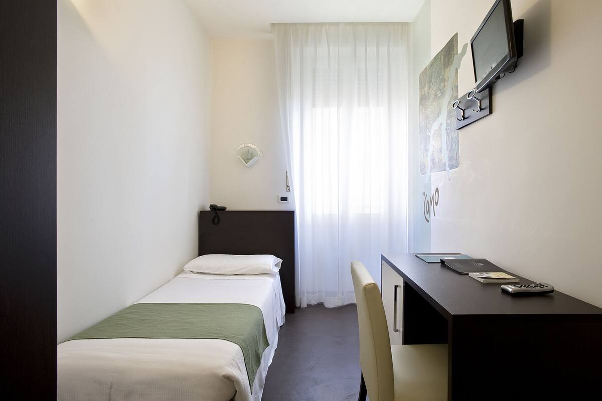 A small hotel room with two beds, a desk, a chair, a TV mounted on the wall, and a window with sheer curtains.