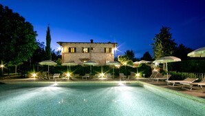 A swimming pool with lounge chairs and umbrellas, surrounded by trees and a stone building at night. - Il Colombaio Winery and Rooms (Monteriggioni)