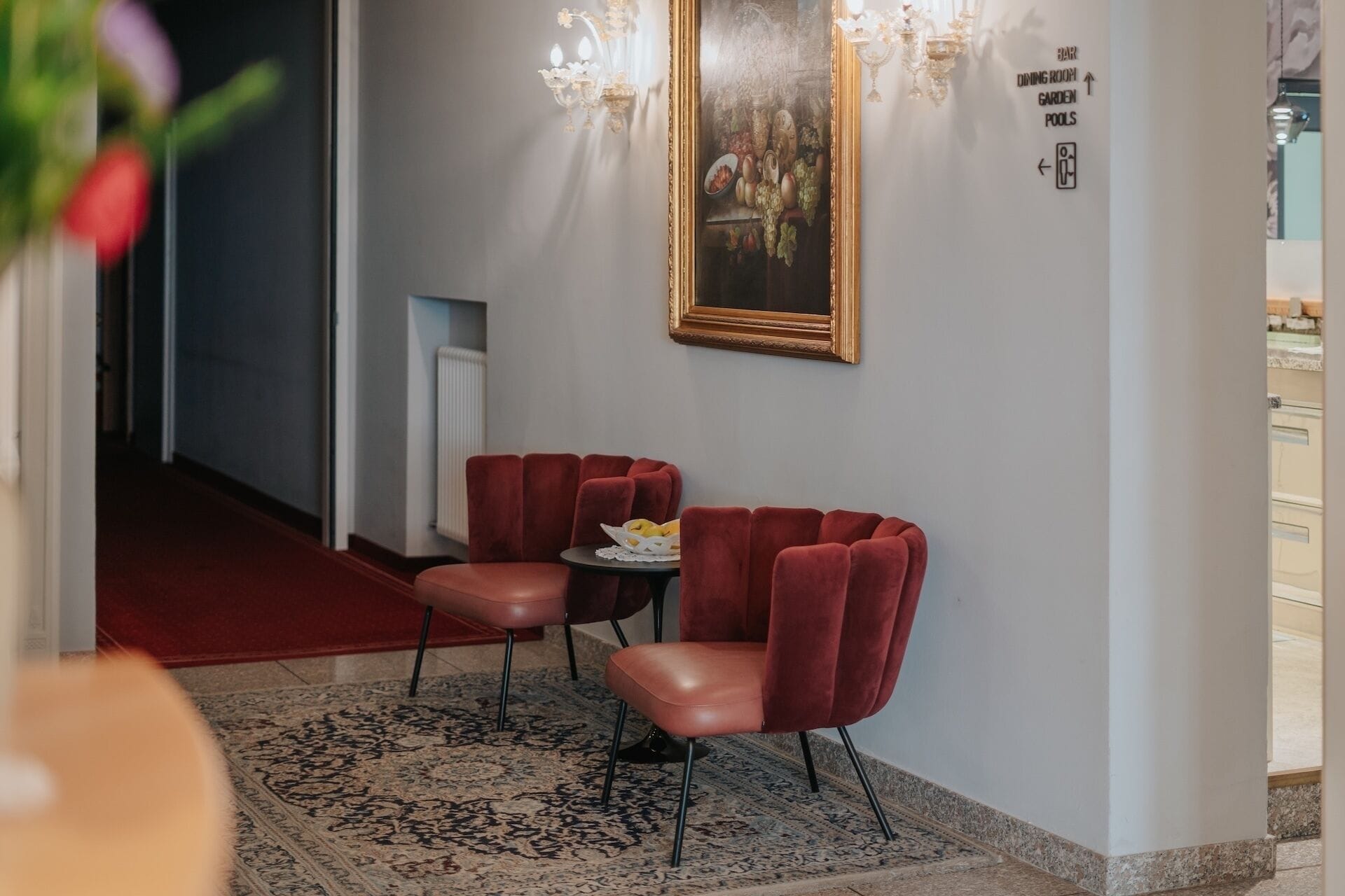 Un pasillo con alfombra roja, dos sillones de terciopelo rojo, una mesa pequeña con una cesta, una araña y un cartel que indica bar, comedor, jardín y piscinas.