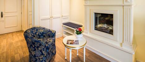 Una habitación con un sillón azul estampado con flores, una mesita blanca con una revista y una chimenea blanca.