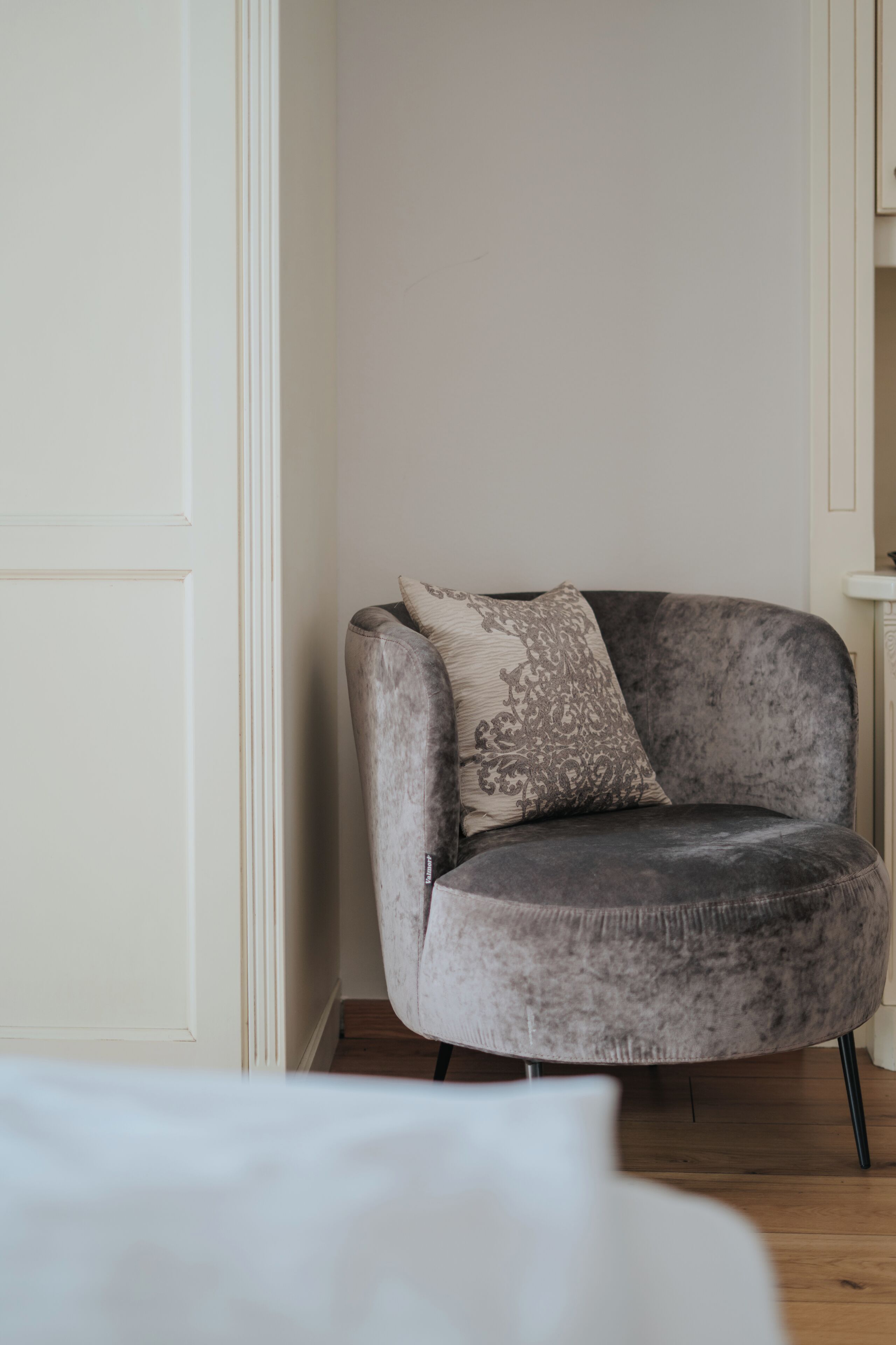 A grey upholstered chair with a patterned pillow in a room with white walls and wooden flooring.