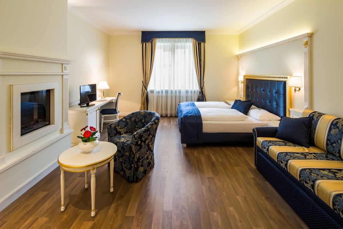 A hotel room with a bed, sofa, armchair, TV, and a small table with flowers.