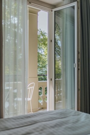 A room with a bed, a window with curtains, and a view of a balcony with a chair and a table.