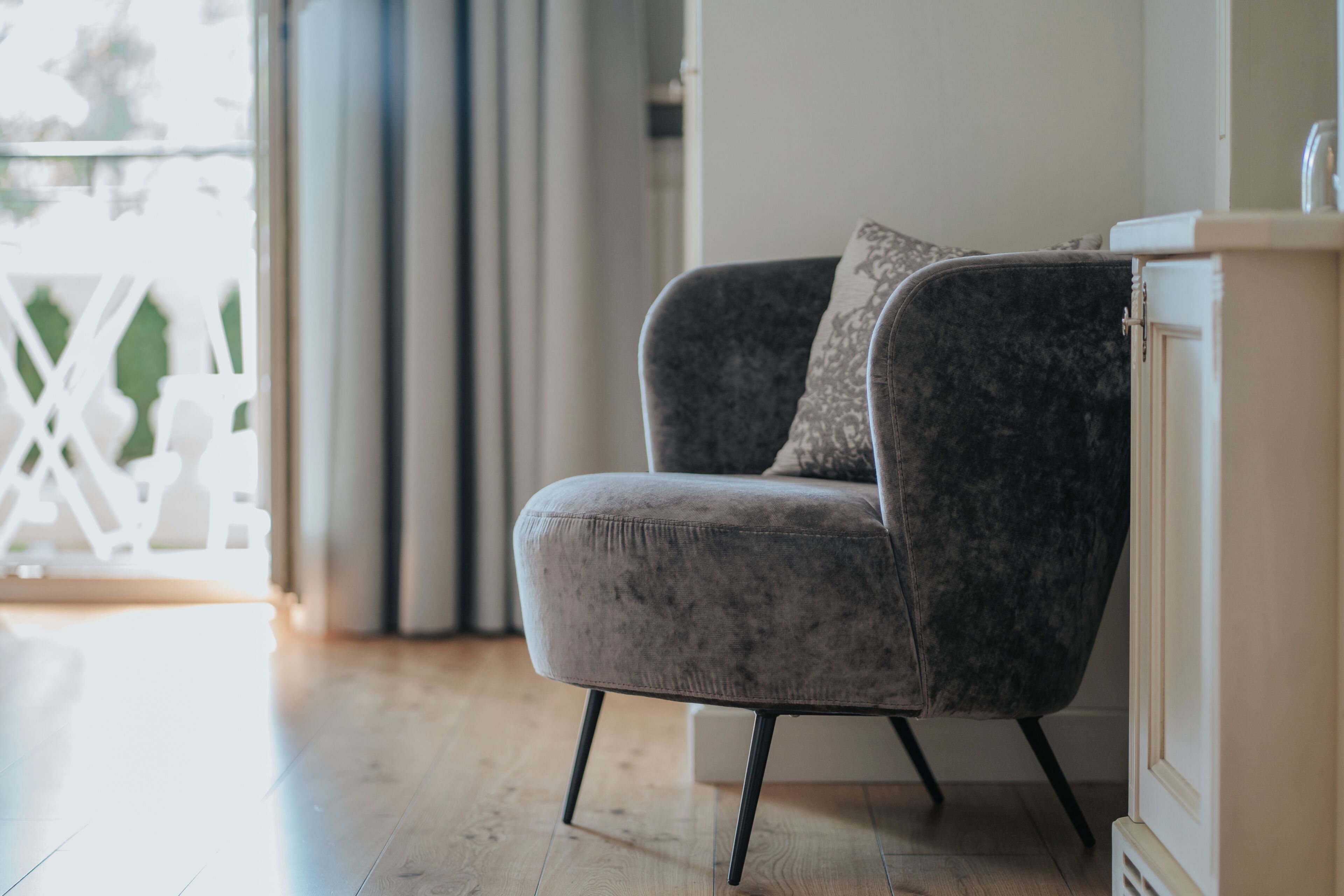 Un fauteuil gris rembourré avec un coussin, placé à côté d’une armoire blanche dotée d’un miroir, dans une pièce au plancher en bois et avec une rambarde blanche en arrière-plan.