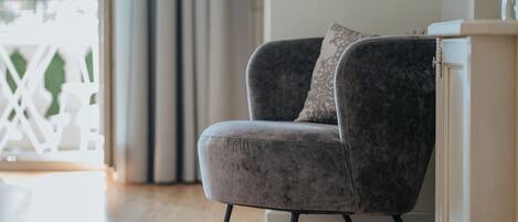 A grey upholstered armchair with a cushion, positioned next to a white cabinet with a mirror, in a room with wooden flooring and a white railing in the background.