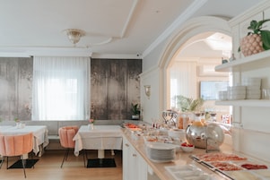 A dining area with tables, chairs, a buffet with food and drinks, and a kitchen area with shelves.