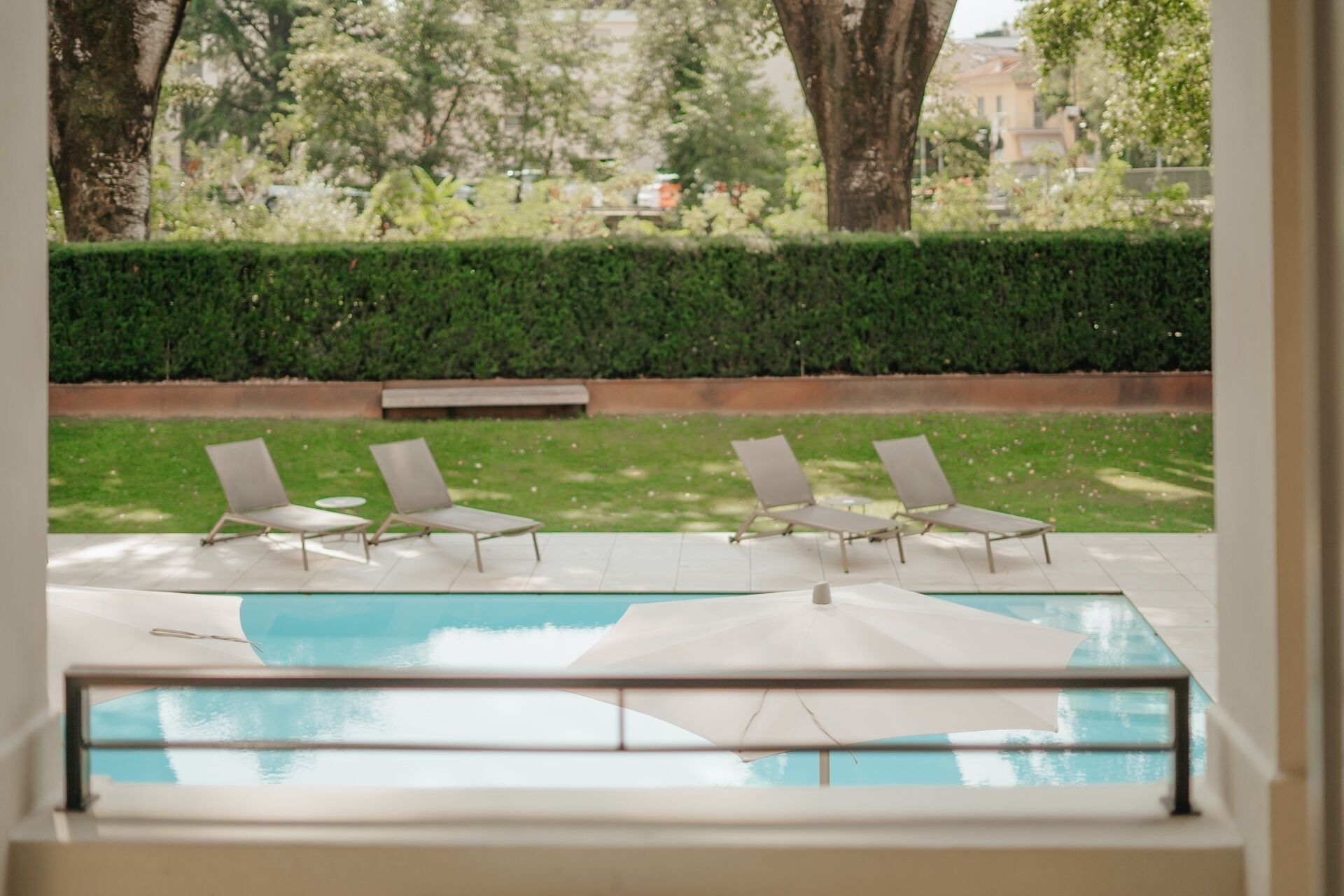 A pool area with lounge chairs and umbrellas, surrounded by greenery and a hedge.