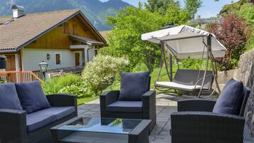 Un’area salotto all'aperto con mobili in vimini e una poltrona a dondolo, con vista su montagne e una casa.
