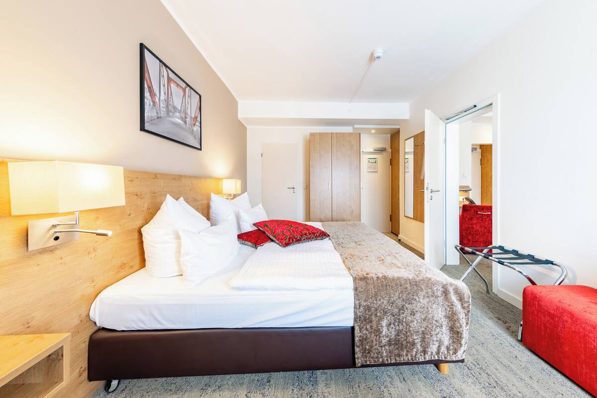 Een hotelkamer met een bed, nachtlampjes, een houten hoofdbord, een rode bank en een deur die naar een andere kamer leidt.