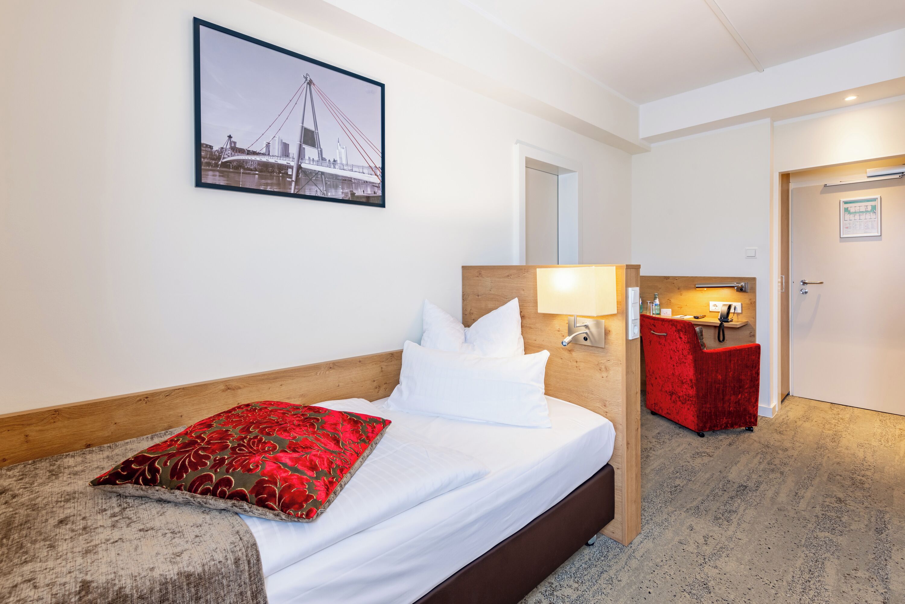 A hotel room with a bed, a red chair, a desk with a lamp, and a framed picture on the wall.