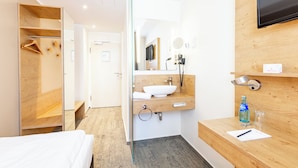 Een moderne hotelkamer met een houten garderobe, een glazen douchecabine, een houten wastafel met een wasbak en een flatscreen televisie aan de muur.