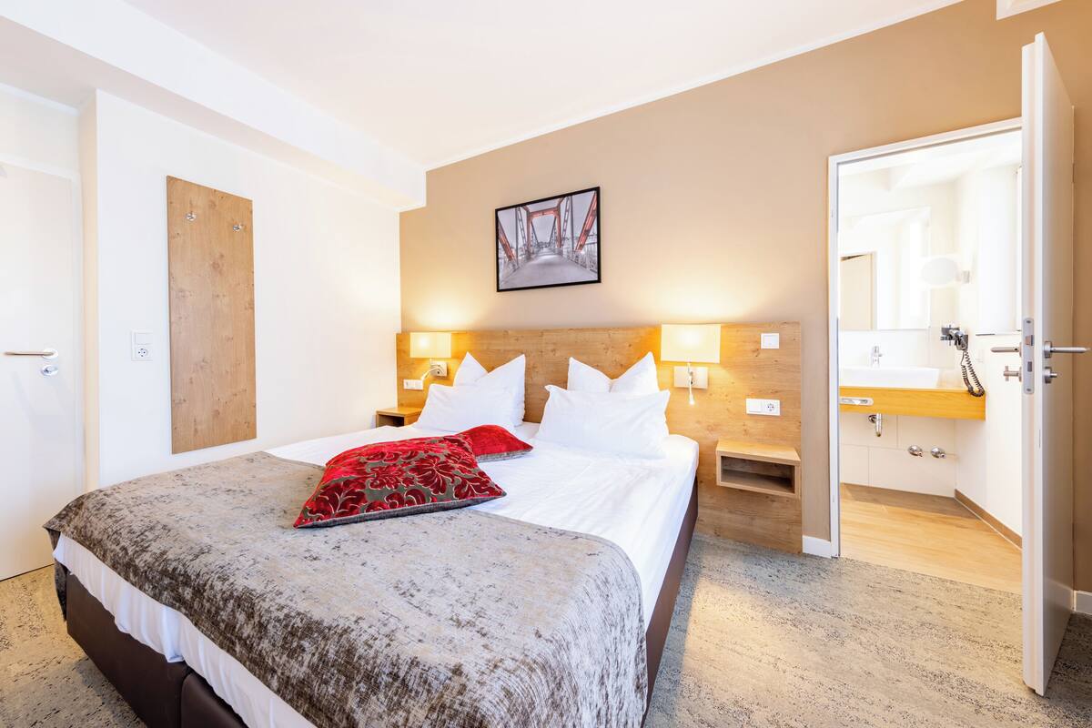 Een hotelkamer met een bed, een houten hoofdbord, twee bedlampen, een rode sierkussen, een ingelijst foto aan de muur, en een open deur naar de badkamer.