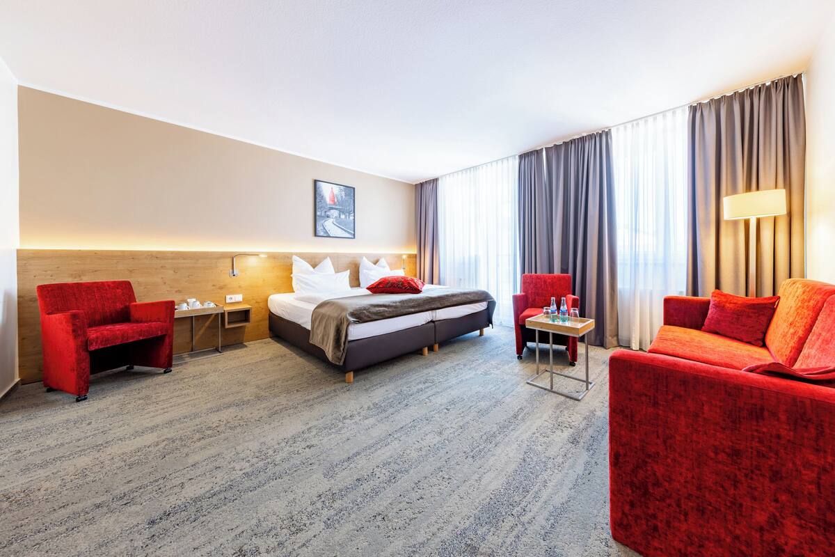 Een hotelkamer met een bed, twee rode fauteuils, een klein tafeltje met een lamp en een groot raam met gordijnen.
