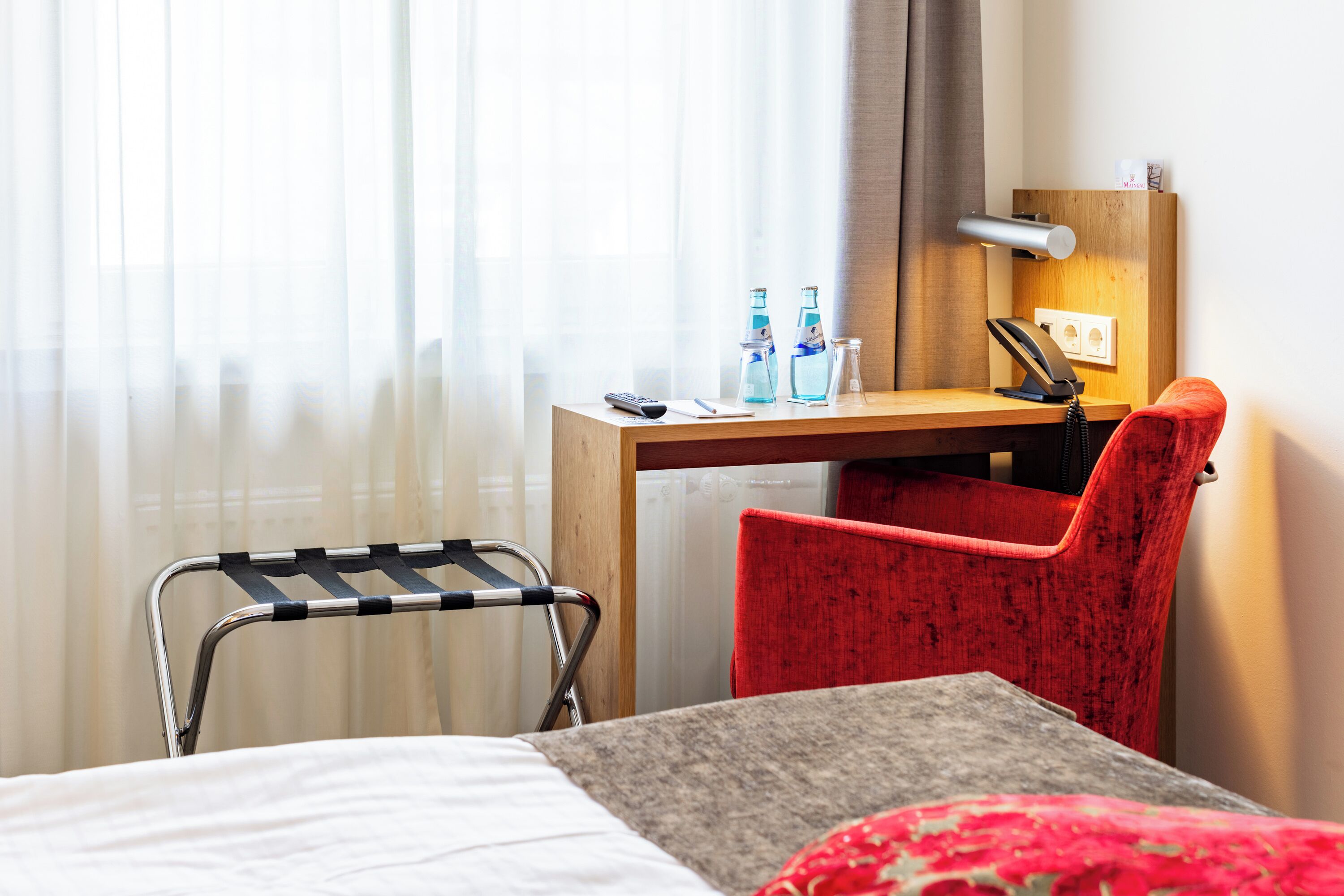 A hotel room with a bed, a desk with a chair, a bedside table, a lamp, and a window with sheer curtains.