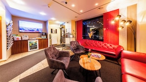 Een moderne hotellobby met een rode bank, twee fauteuils, een tafeltje met een vaas, een aan de muur bevestigde televisie, een receptiebalie en een koelkast.