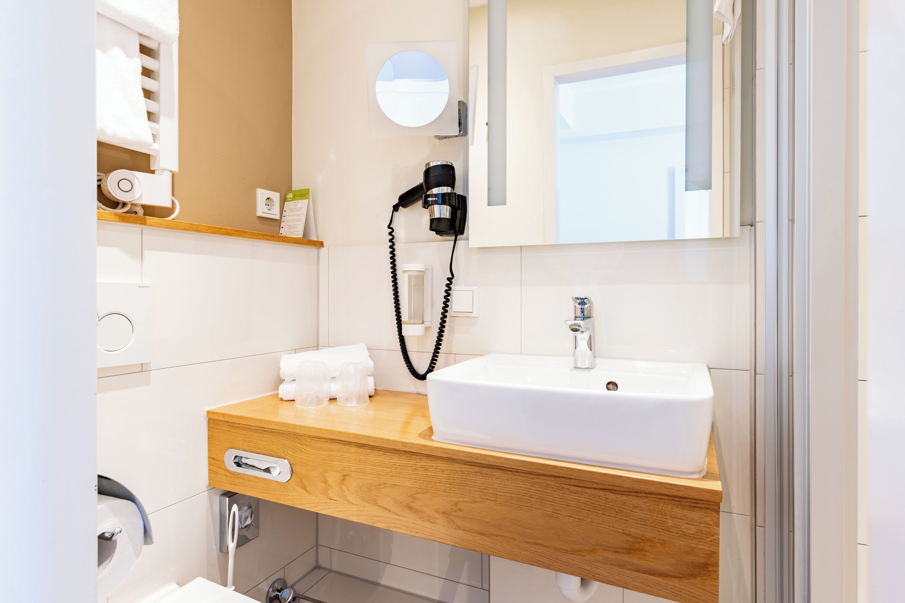 A modern bathroom with a wooden vanity, white sink, and a hairdryer on the wall.