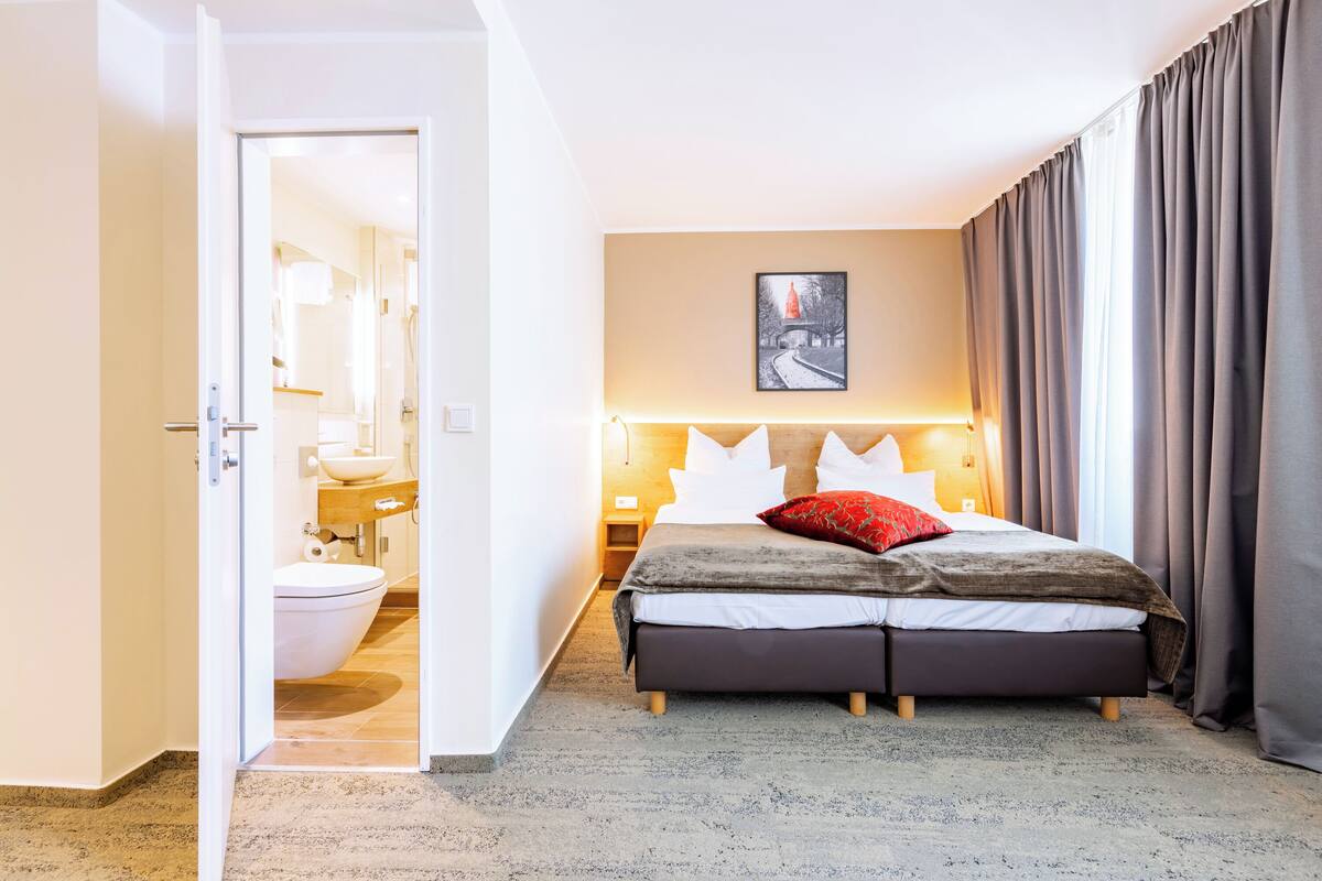 Hotelkamer met een bed, nachtkastje, raam met gordijnen, een badkamer zichtbaar door een geopende deur en een ingelijst schilderij aan de muur.