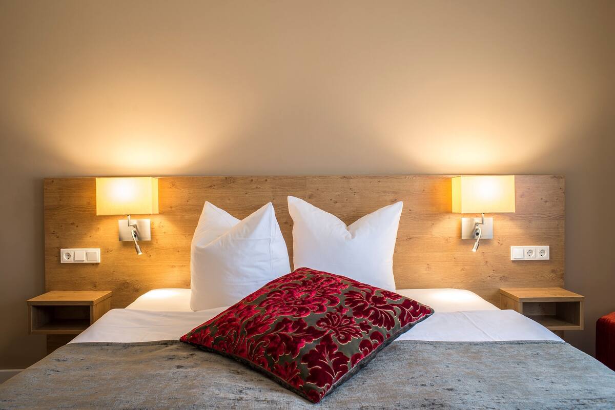 Hotelkamer met houten hoofdeinde, twee bedlampen, een bed met witte lakens en een rood kussen met bloemen.