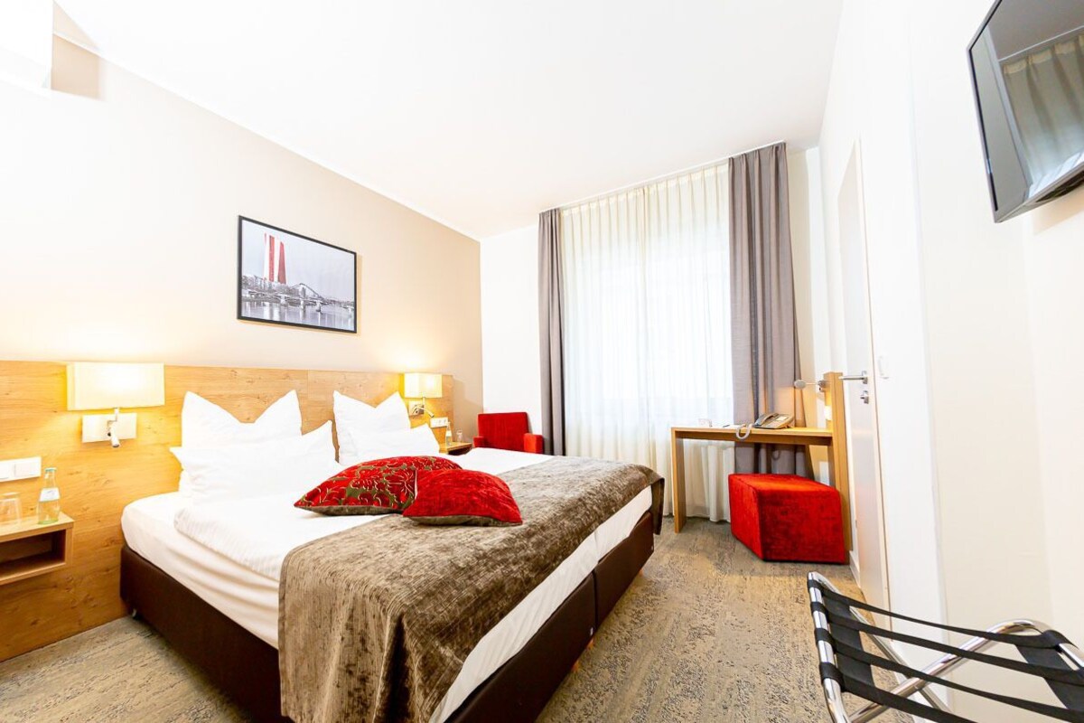 Habitación de hotel con cama, mesitas de noche, escritorio, silla, televisor y ventana con cortinas.