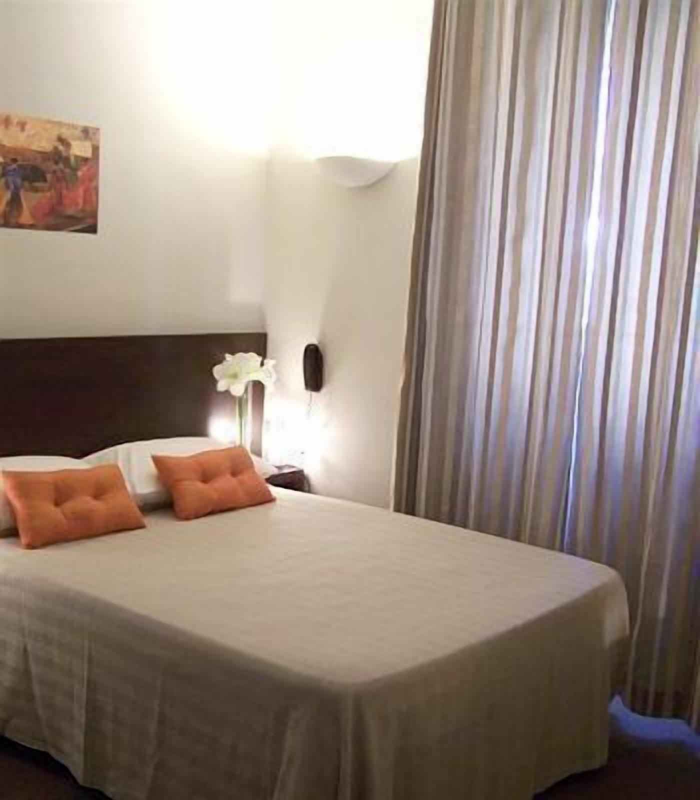 Un letto rifatto con due cuscini arancioni, una parete bianca con un quadro, un comodino con una lampada e una finestra con tende.