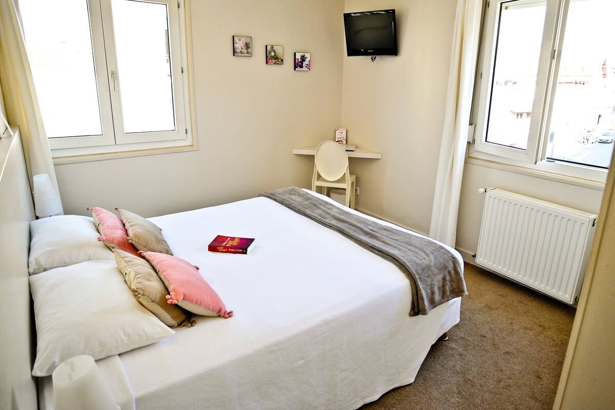 A bedroom with a bed, pillows, a desk, a chair, a TV, and windows.