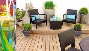 Um deck de madeira com duas cadeiras de vime preto, uma mesa pequena e vasos com plantas.