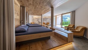 A modern bedroom with a wooden ceiling, a large bed with blue bedding, a wooden desk, and a view of the outdoors. -  Hotel Platz (Castelrotto)