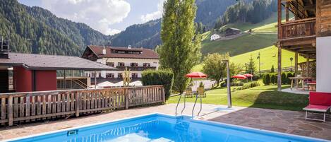 A swimming pool with a wooden deck, a red building, and a mountain backdrop.