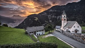 A church with a clock tower, surrounded by a rural landscape with mountains in the background. -  Hotel Platz (Castelrotto)