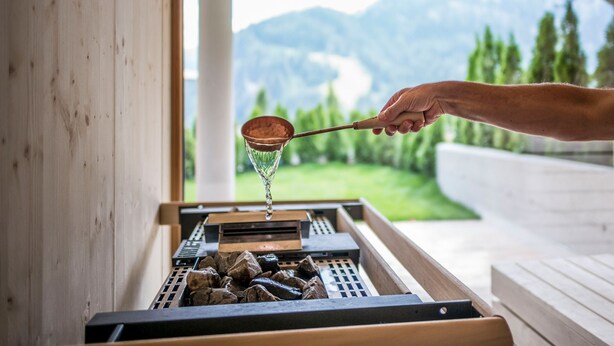 A hand using a copper ladle to pour water onto a grill with rocks. -  Hotel Platz (Castelrotto)