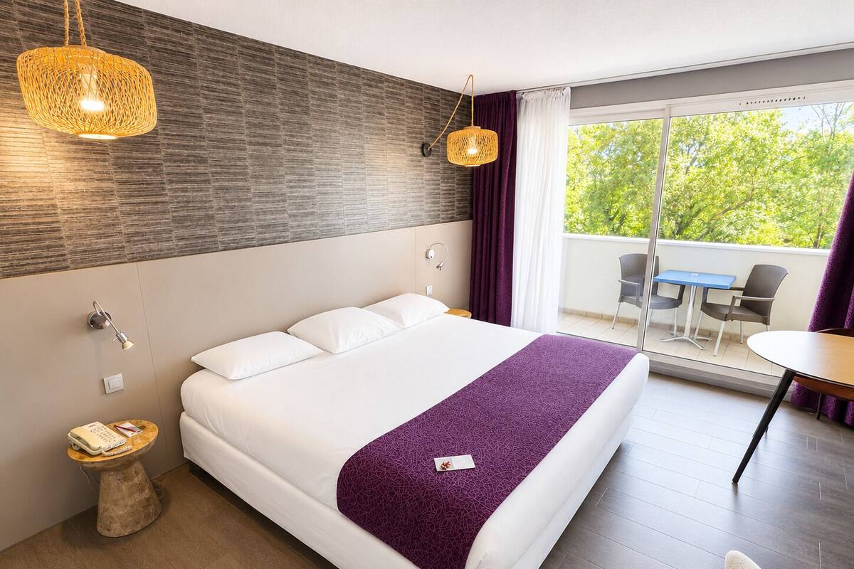 Hotelkamer met een bed, een klein nachtkastje met telefoon, een wandlamp, een raam met gordijnen en een balkon met stoelen en een tafel.