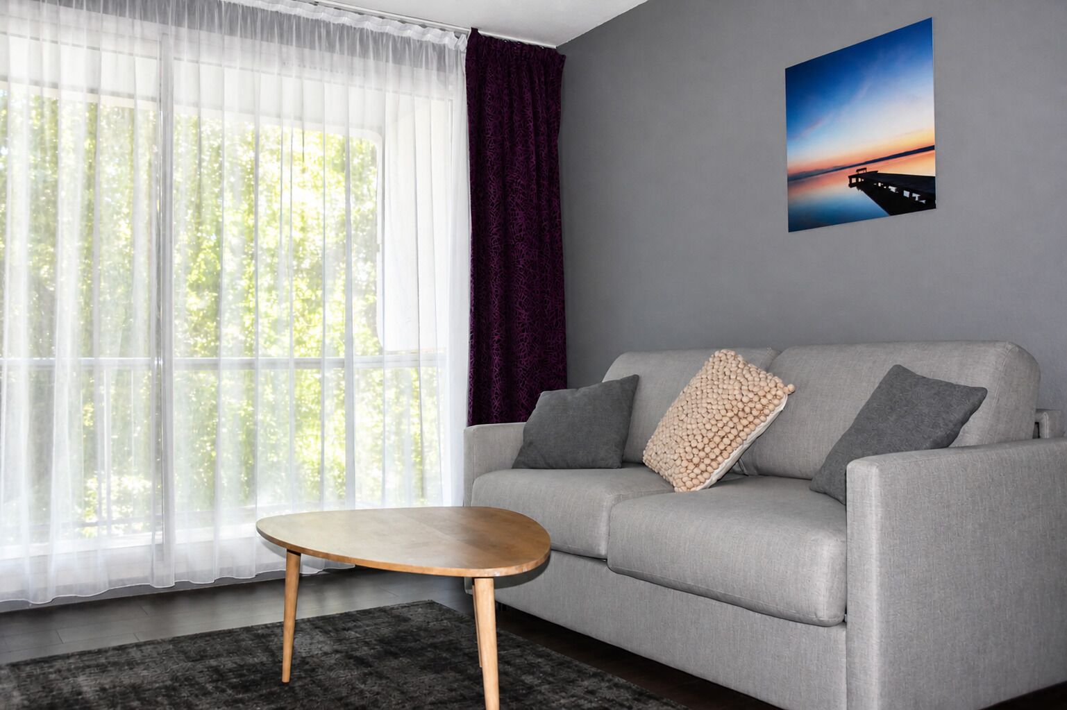 Una sala de estar moderna con un sofá gris, una mesa de centro redonda de madera, cortinas blancas translúcidas, una cortina morada y un cuadro en la pared.