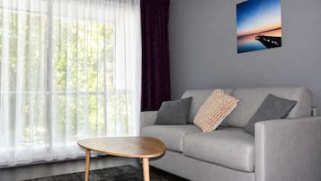 Un salon moderne avec un canapé gris, une table basse ronde en bois, des rideaux blancs transparents, un rideau violet et un tableau encadré accroché au mur.