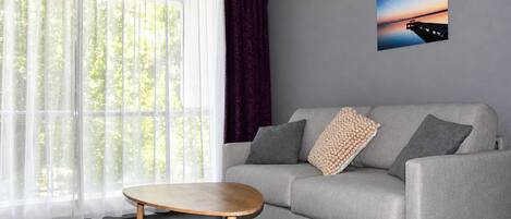 Una sala de estar moderna con un sofá gris, una mesa de centro redonda de madera, cortinas blancas translúcidas, una cortina morada y un cuadro en la pared.