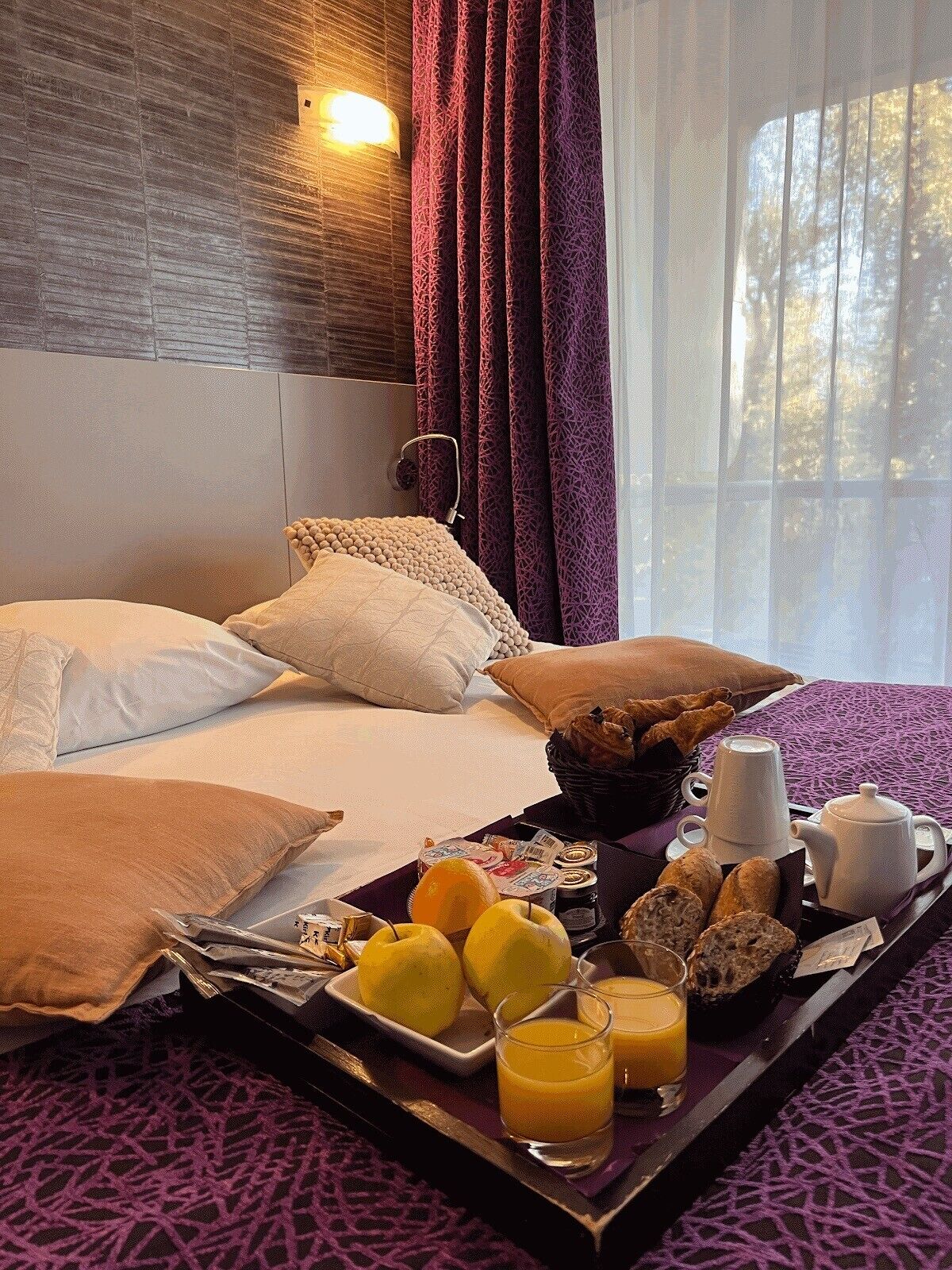 A hotel room with a bed, pillows, a tray with breakfast items, and a window with curtains.
