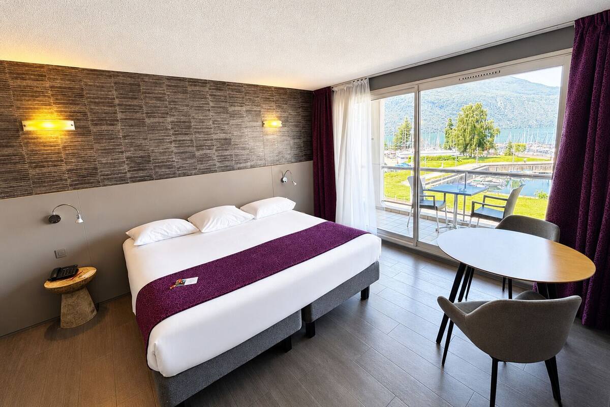 Een hotelkamer met een bed, nachtkastje, lamp en uitzicht op een jachthaven.