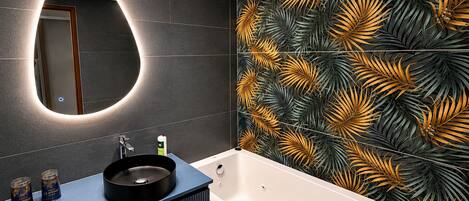 Un baño moderno con un espejo redondo, una bañera y un tocador con encimera oscura.