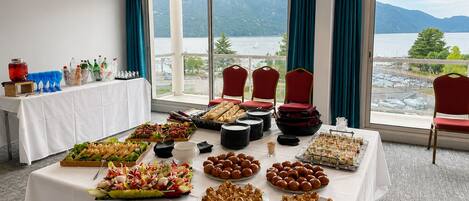 Una mesa de buffet con diferentes alimentos, una vista de un lago y montañas a través de amplios ventanales, y sillas rojas.