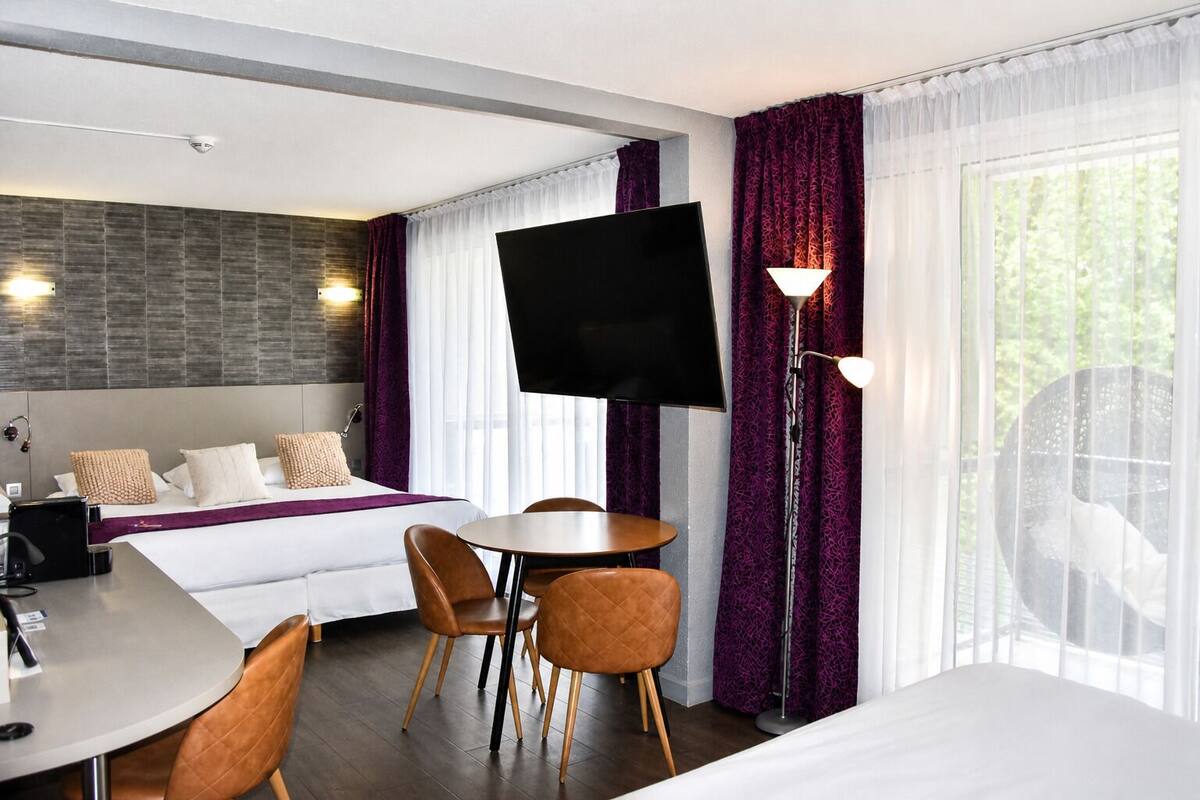 Een hotelkamer met een bed, een bureau, twee stoelen, een televisie en een groot raam met gordijnen.