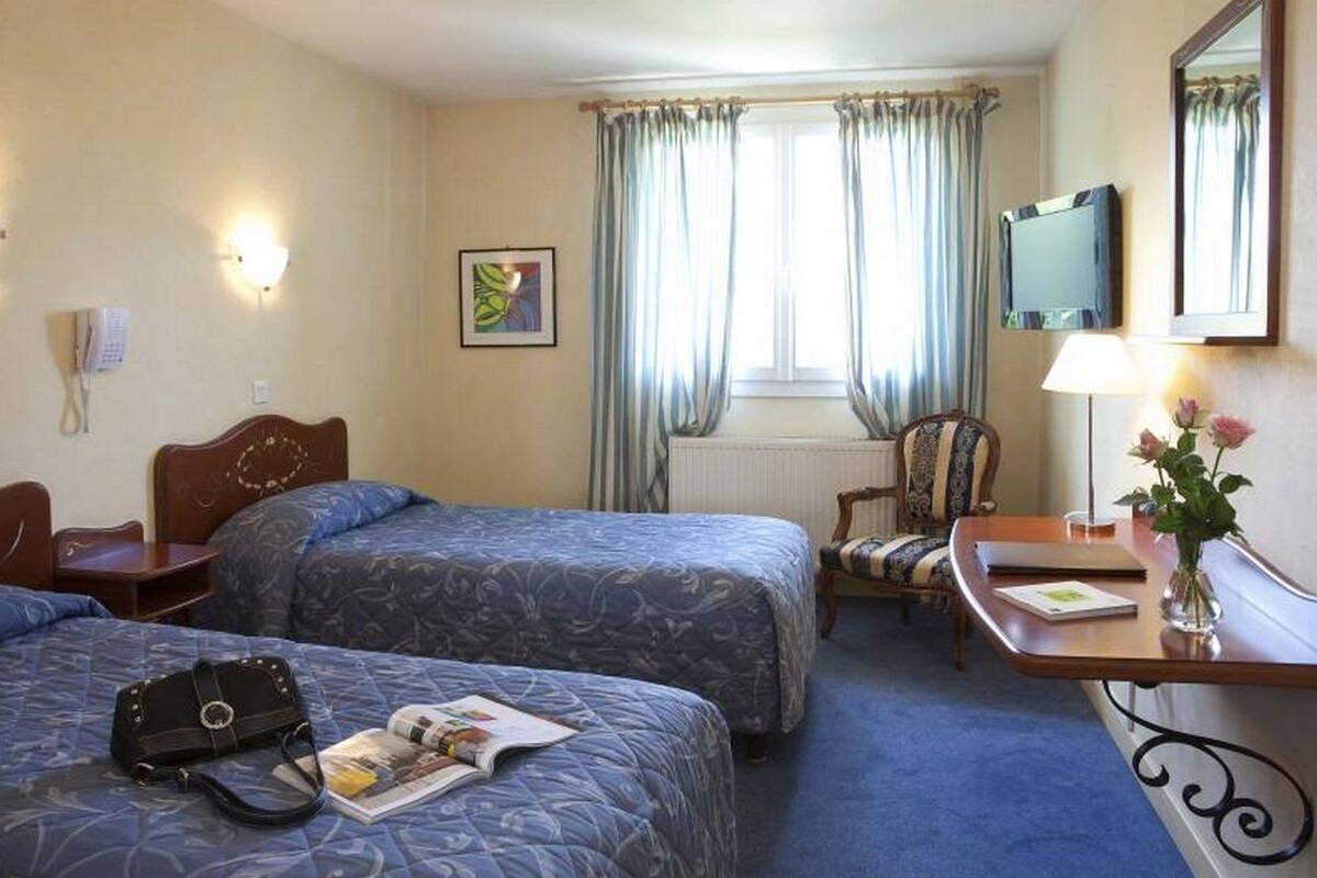 Habitación de hotel con dos camas, un escritorio, una silla, un televisor, una lámpara y una ventana con cortinas.