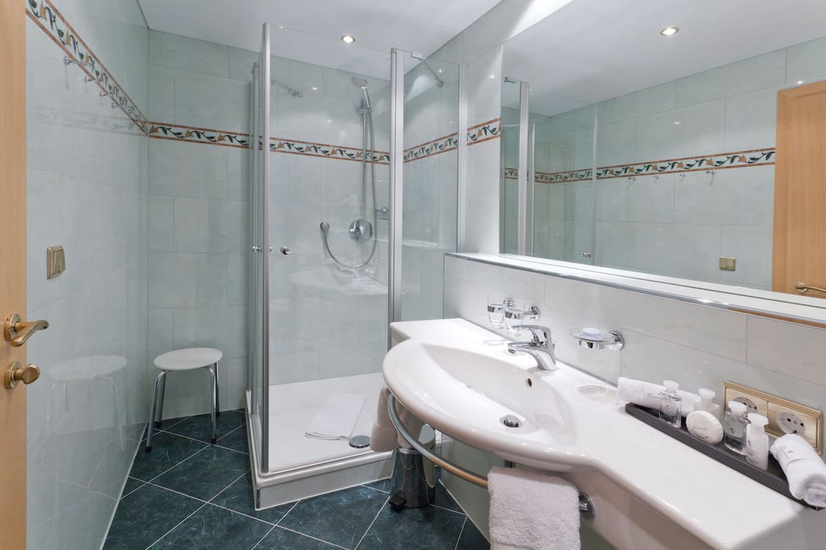 Un baño moderno con ducha de vidrio, lavamanos blanco y artículos de aseo, además de un taburete pequeño.