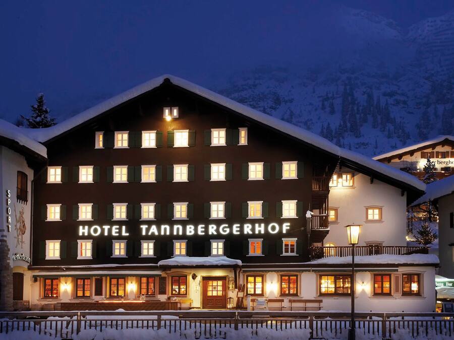 A hotel with a sign reading "HOTEL TANNBERGERHOF" in front of a snowy mountain backdrop.