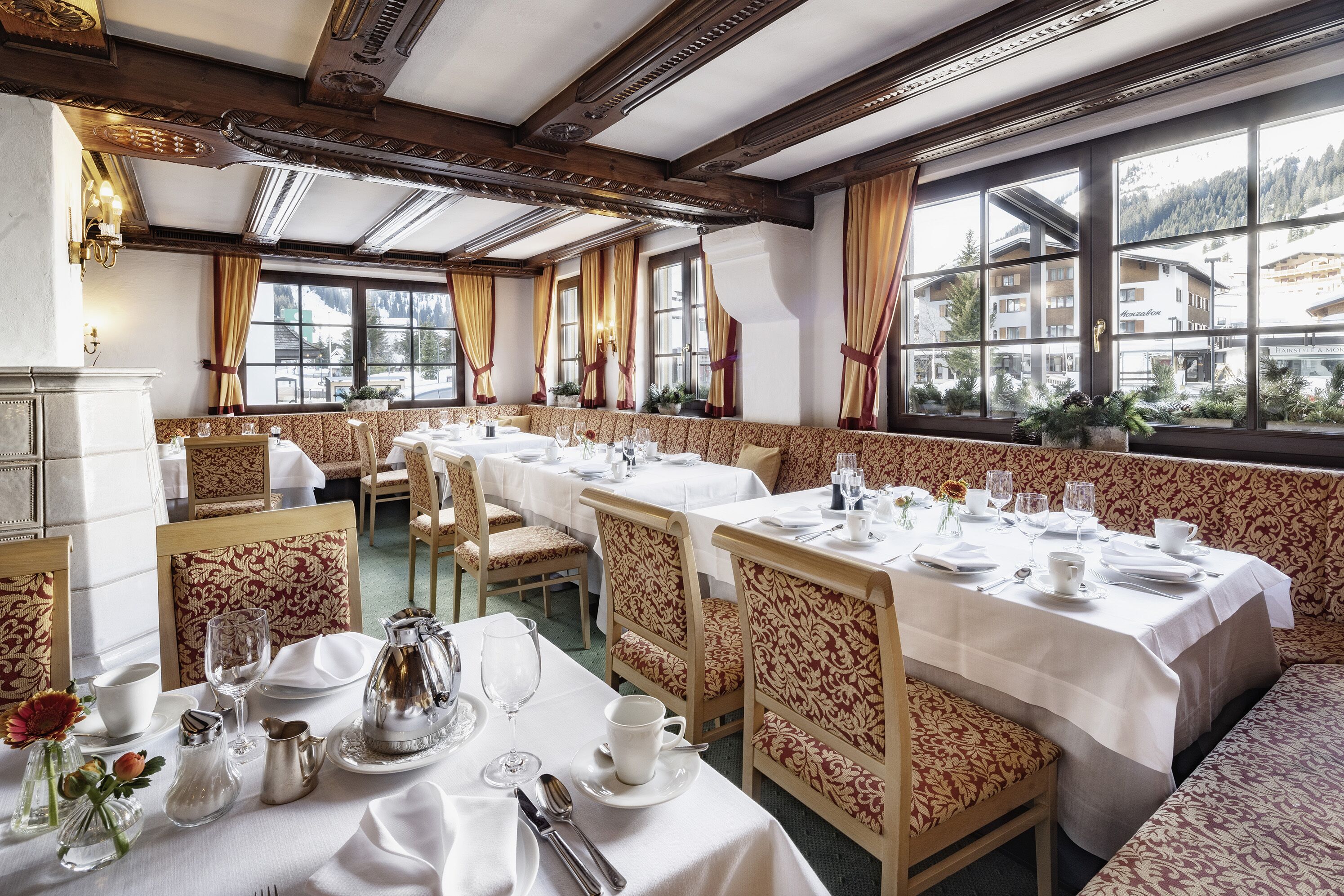 Uma sala de jantar com mesas longas preparadas para vários convidados, cadeiras de madeira com estofados estampados e grandes janelas com vista para uma paisagem montanhosa.