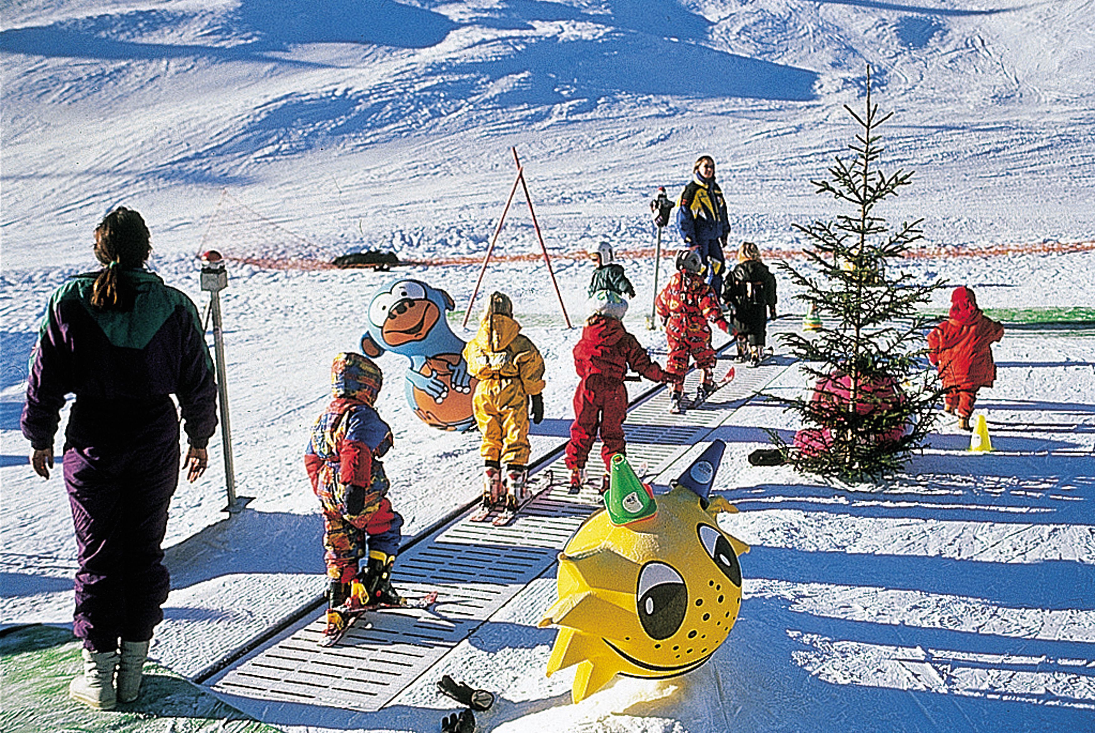 A snowy outdoor scene with people in winter clothing, a small tree, and a large yellow snowman figure.