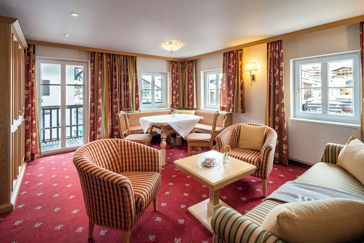 Habitación de hotel con alfombra roja, sillones a rayas, un sofá, una mesa de comedor y vistas a edificios a través de grandes ventanales.